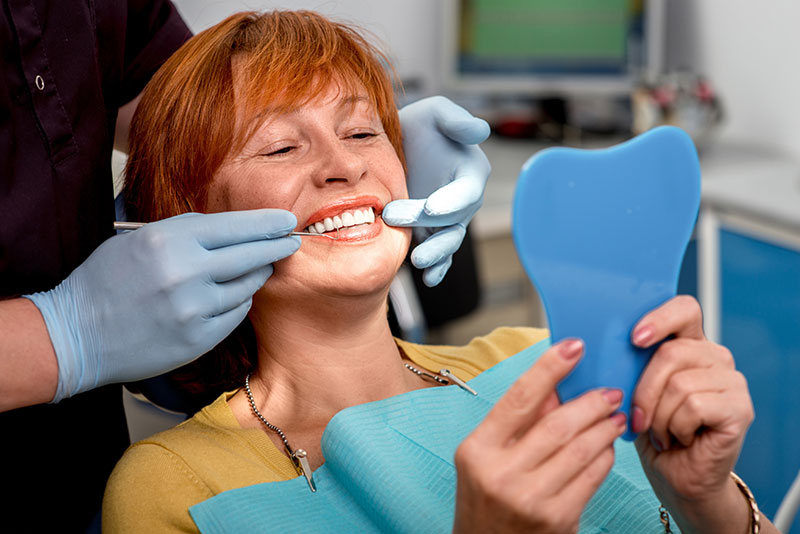 A woman is sitting in a dental chair, smiling broadly while holding a blue toothbrush and looking at her reflection in a mirror. She has red hair and is wearing a white shirt with a black necklace.