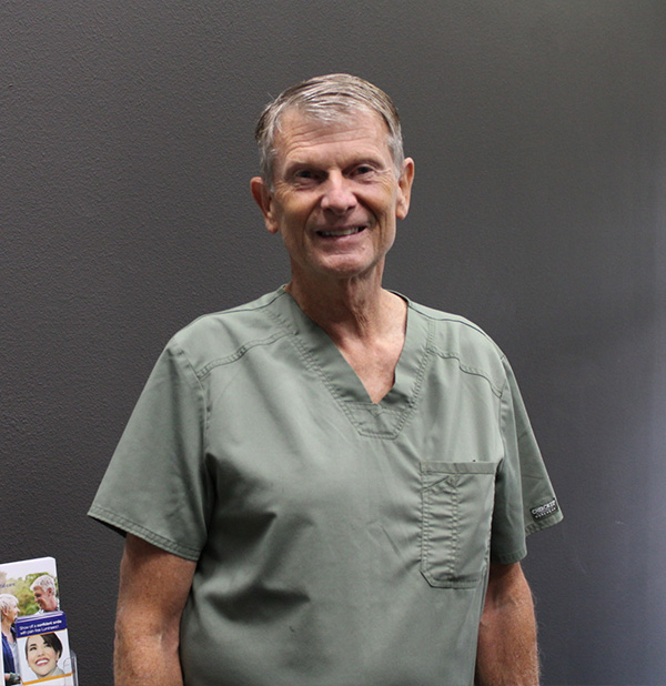 The image shows an older man wearing a light green scrub top, standing in front of a dark background with a cardboard sign to the left.