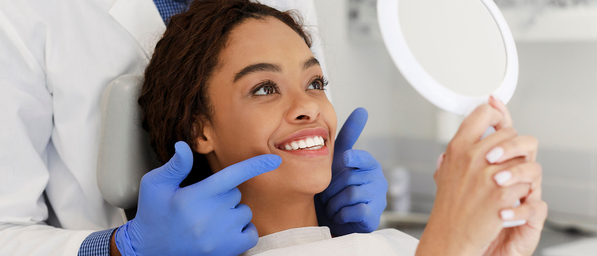 A woman is seated in a dental chair, smiling at the camera with her eyes closed, while a dentist inspects her teeth.