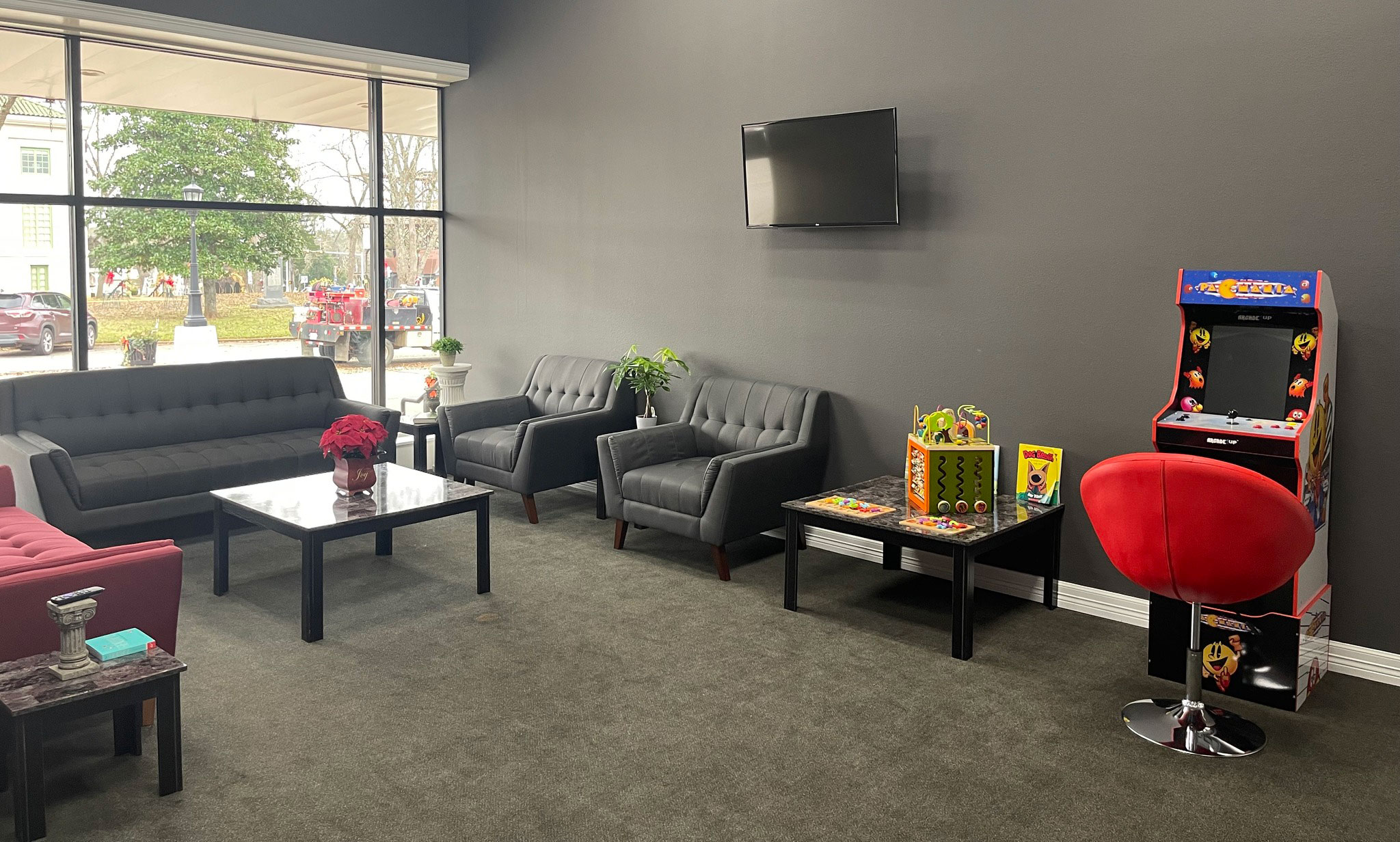 The image shows an interior space that appears to be a waiting or reception area, featuring a modern couch, a red chair with a gaming console, a coffee table, and a wall-mounted television.