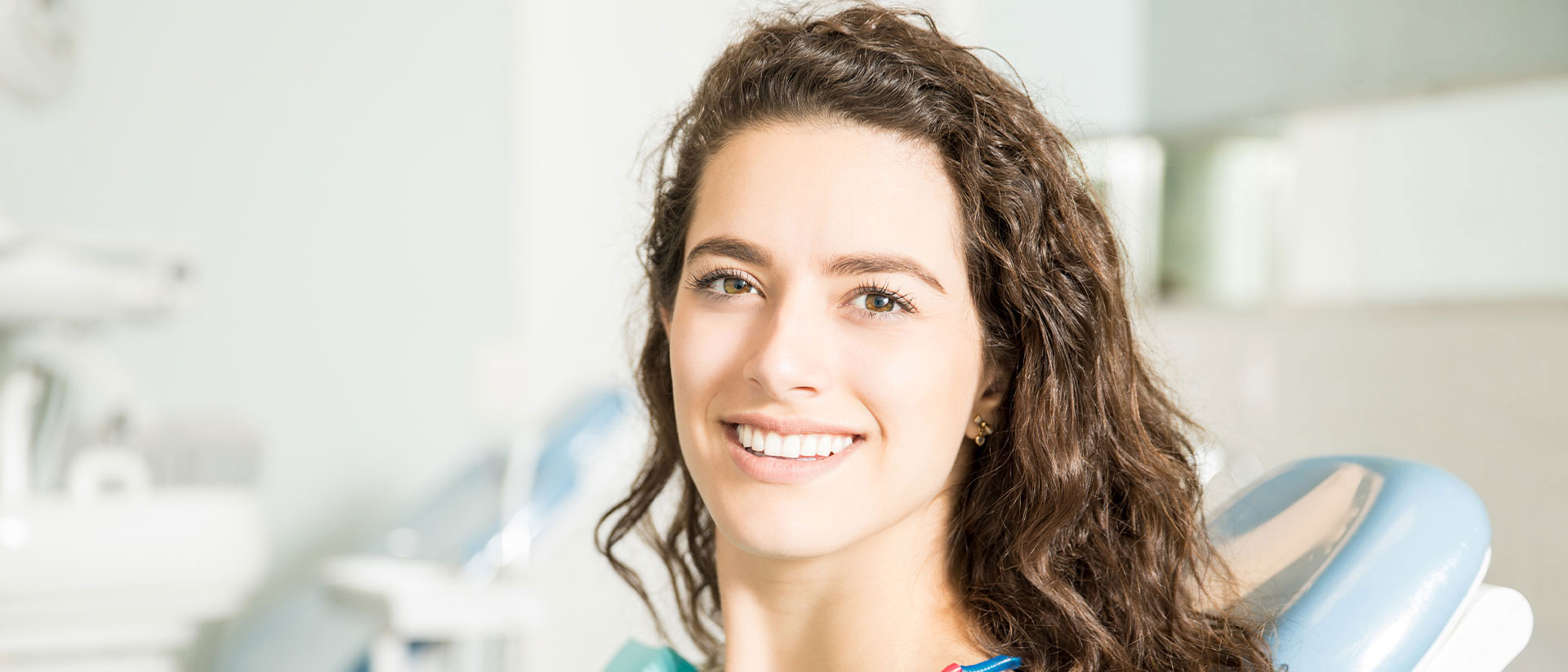 The image is a photograph of a woman sitting in a dental chair, smiling towards the camera. She appears to be at a dental clinic, as indicated by the equipment and setting.