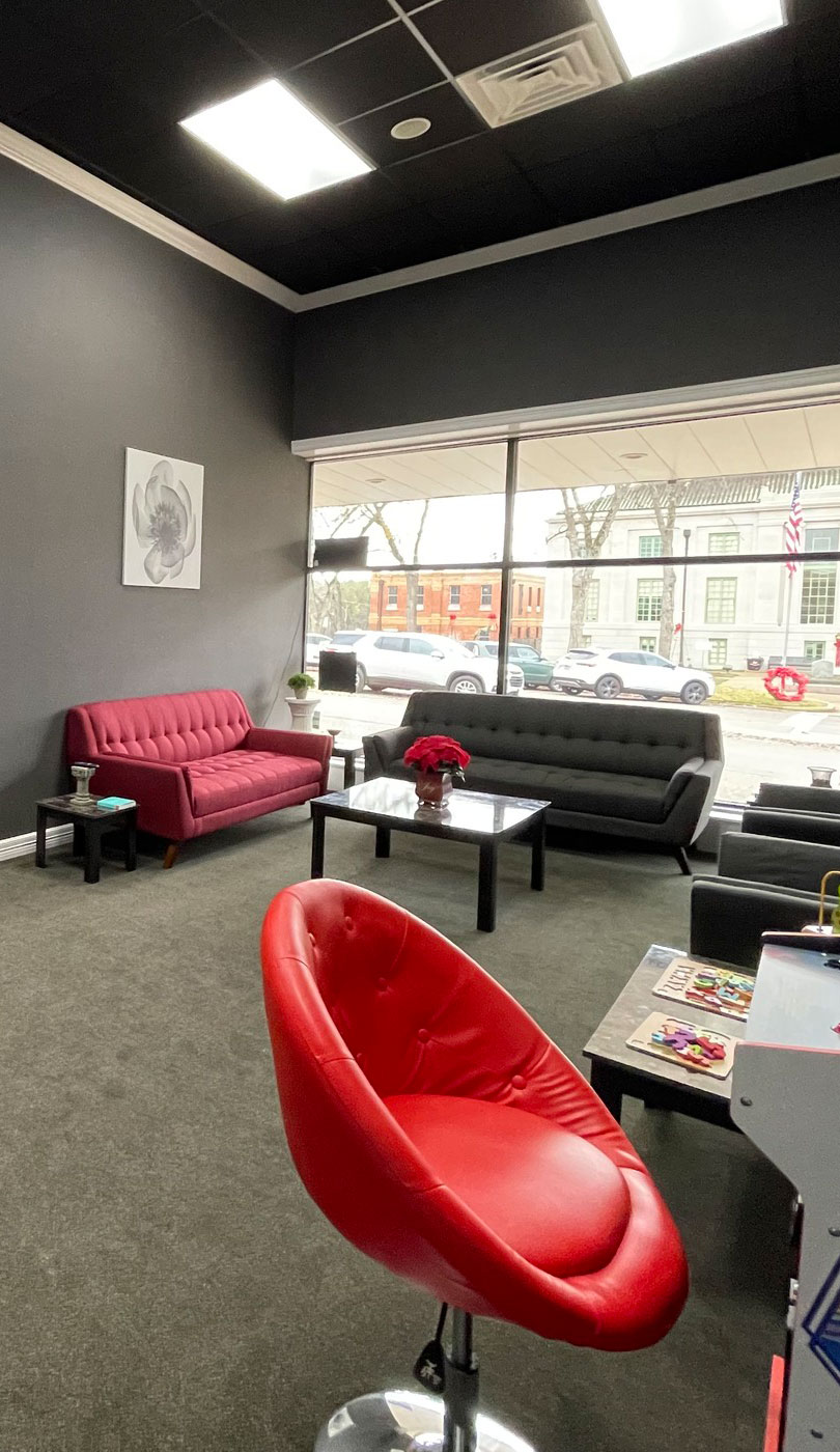 The image shows an interior space that appears to be a modern office or waiting area, featuring a dark room with a red leather chair and ottoman in the foreground, a black carpeted floor, a gray wall, and a large window allowing natural light into the room. There is a black counter with a red stool on one side, and a coffee table with a magazine rack in the center of the space. The office has a sleek design, with minimal decoration and a focus on functionality.