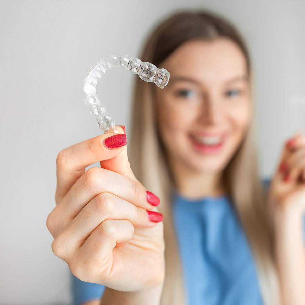 A woman is holding a clear plastic retainer with her right hand, smiling and showing it to the camera.