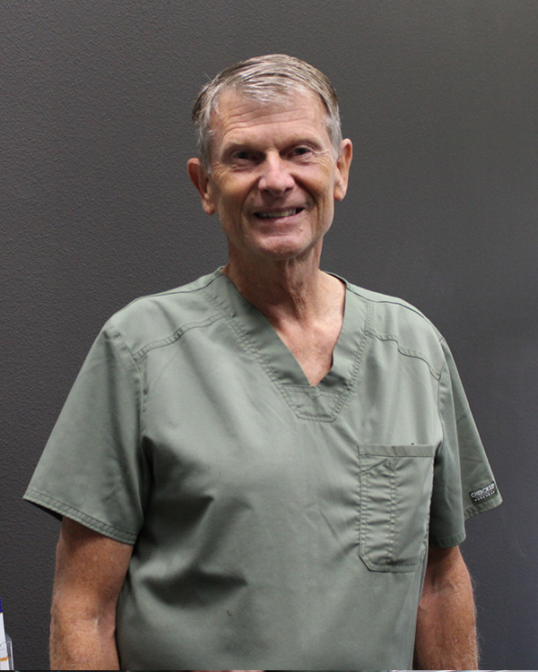 The image shows an older man wearing a green scrub top, standing against a dark background with a neutral expression.