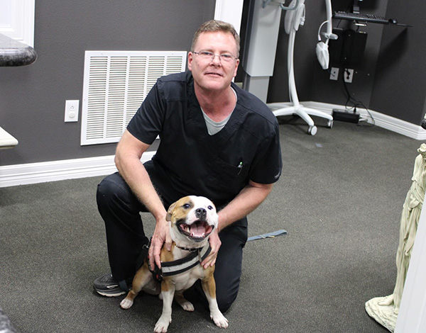 A man in a surgical mask is kneeling next to a brown and white dog with its tongue out, both are indoors.