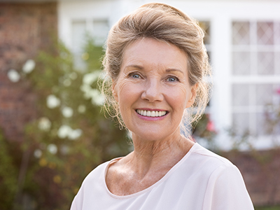 A smiling older woman poses in front of a house with a well-maintained garden.