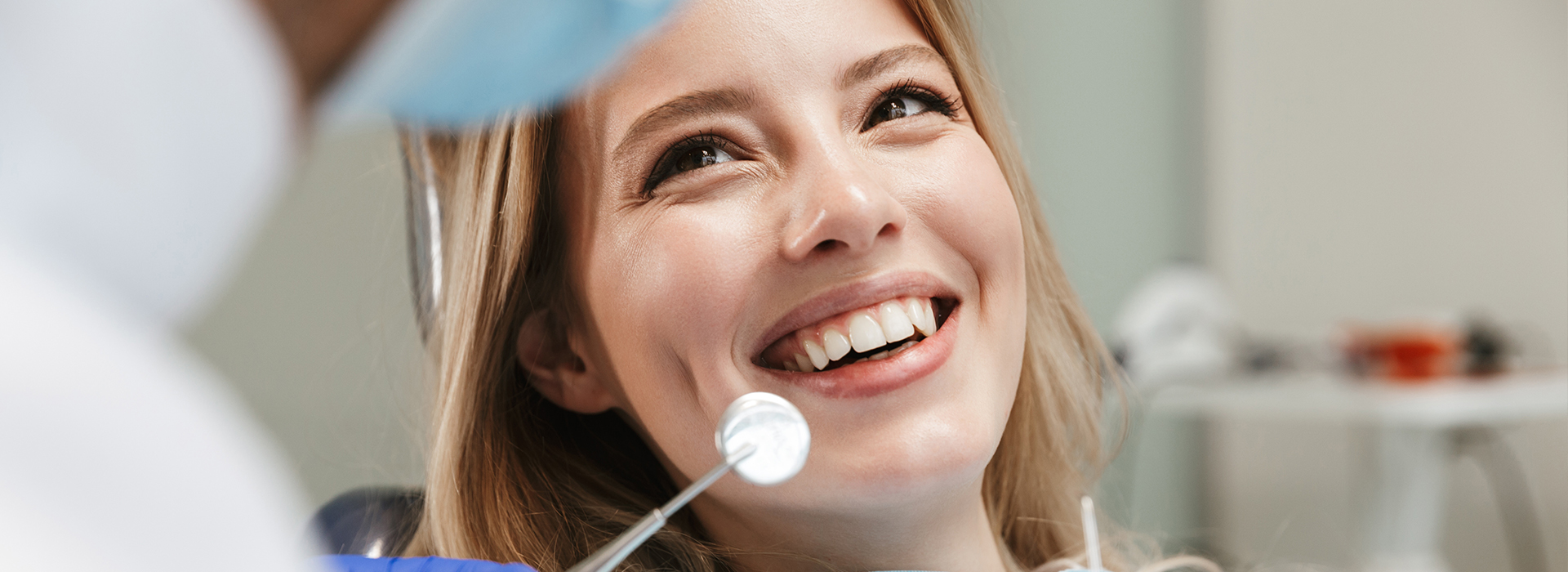 The image shows a woman sitting in front of a dental professional, both wearing masks and looking at the camera with smiles.