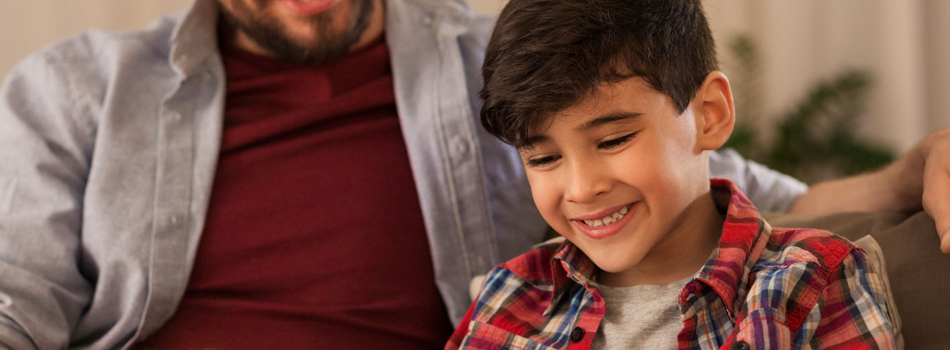 The image shows a man and a young boy sitting closely together, both smiling and looking at something off-camera. They appear to be in a relaxed indoor setting.