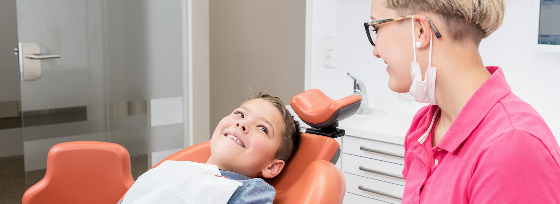 A young child in a dental chair, smiling and looking up at the camera, with a woman in pink standing behind him, likely a dentist or dental assistant.