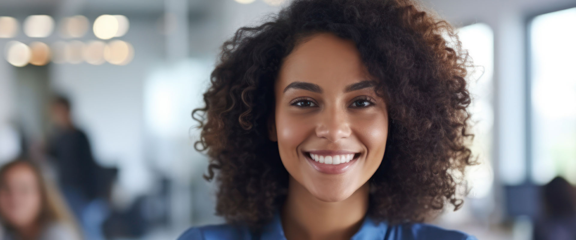 A woman with a radiant smile, wearing a blue shirt and standing in an office environment.