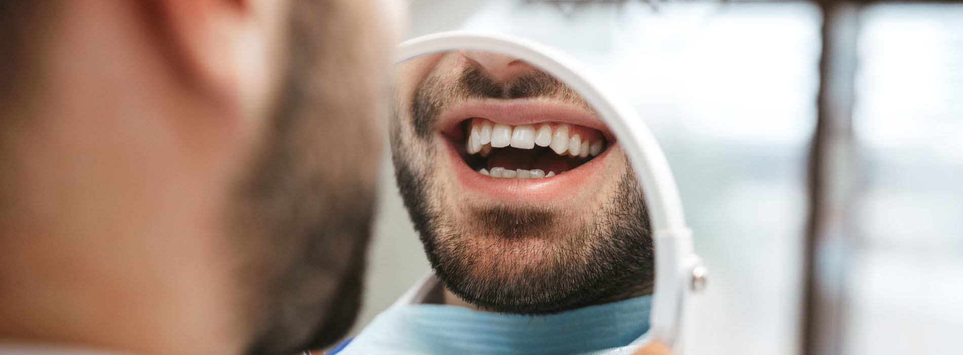 The image shows a close-up of a person with a beard, wearing a blue mask and holding a dental instrument, looking directly at the camera while smiling.