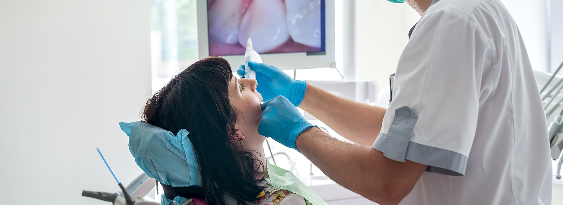 The image shows a dental professional performing a procedure on a patient in a dental office setting, with the patient seated in a chair and wearing a blue surgical mask.