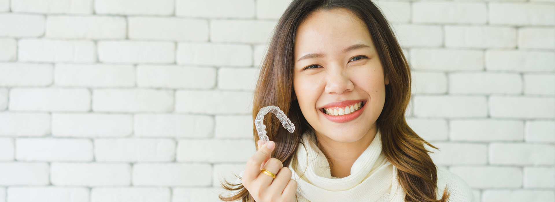 A young woman with a radiant smile is holding a ring, standing in front of a white brick wall.