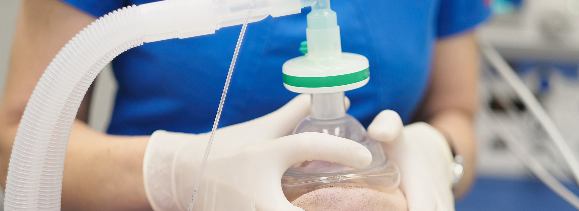 The image shows a person wearing gloves and a blue shirt, holding a clear plastic tube with a green cap connected to a medical device, suggesting a healthcare or laboratory setting.