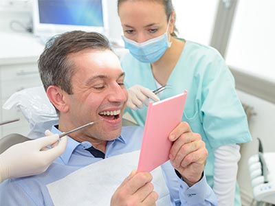 The image depicts a man sitting in a dental chair, holding up a pink card, with a smile on his face. He appears to be engaged in a humorous or lighthearted moment with a female dentist standing behind him, who is looking at the card and seems to be laughing.