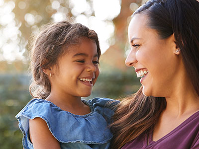 A woman and a child are smiling at the camera. The woman is standing, and the child is seated on her hip. They appear to be outdoors during daylight.