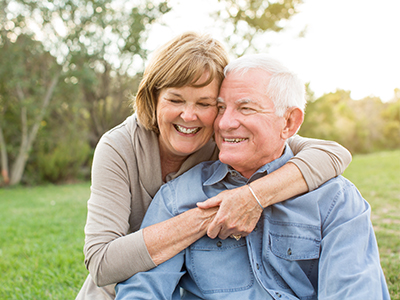 This is a photograph of an elderly couple embracing each other, with the man wearing a blue shirt and the woman in a beige top, set against a blurred background that suggests an outdoor setting.