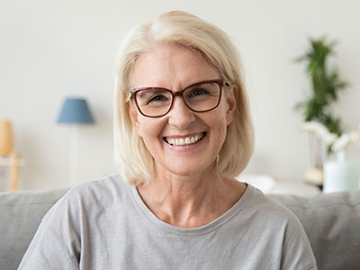 The image features a smiling woman with short blonde hair, wearing glasses and a gray top, sitting in an indoor setting.