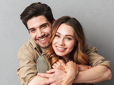 The image is a photograph of a man and a woman embracing each other. They appear to be in their late twenties or early thirties, with the man on the left wearing a light brown shirt and the woman on the right wearing a dark top. Both individuals are smiling and looking at the camera. The background is neutral and does not distract from the subjects.