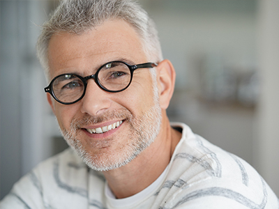 The image shows a man with gray hair, wearing glasses and a white shirt, smiling slightly at the camera. He appears to be middle-aged or older, and he has a beard.