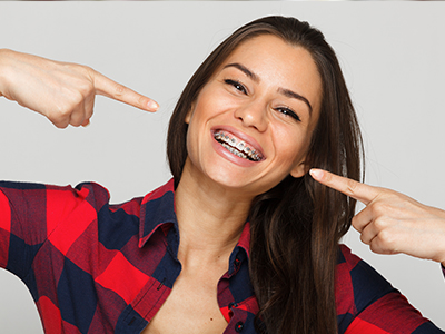 The image is a photograph of a woman with a wide smile, pointing towards her teeth with her right hand. She has long hair, wears plaid flannel and has a playful expression on her face.