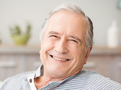The image shows an elderly man with gray hair, wearing a blue shirt, sitting in a relaxed posture with a smile on his face.