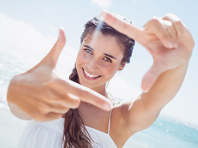 A smiling woman with long hair, holding up a hand in front of her face to create a frame or viewfinder effect.