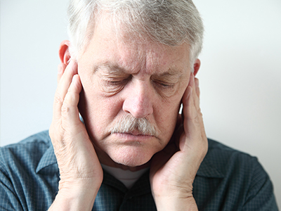 The image depicts an older man with his eyes closed, holding his face with both hands, as if in pain or discomfort. He appears to be indoors and is wearing a blue shirt.